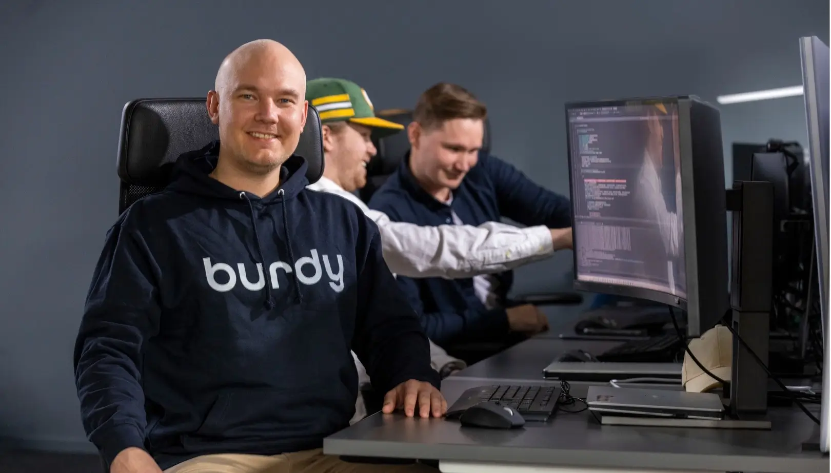 A male employee sits on a computer chair with a hoodie with the text "Burdy" facing the camera. Besides him he has his computer and behind him his two colleagues are point to another computer screen