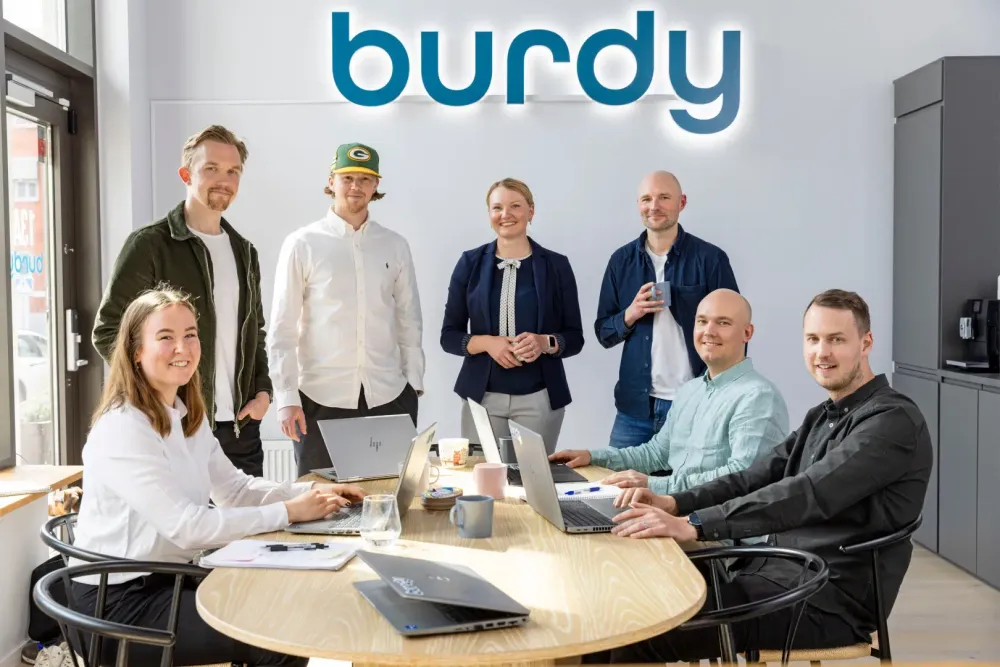 A team (7 people) is sitting and standing around a table looking at the camera