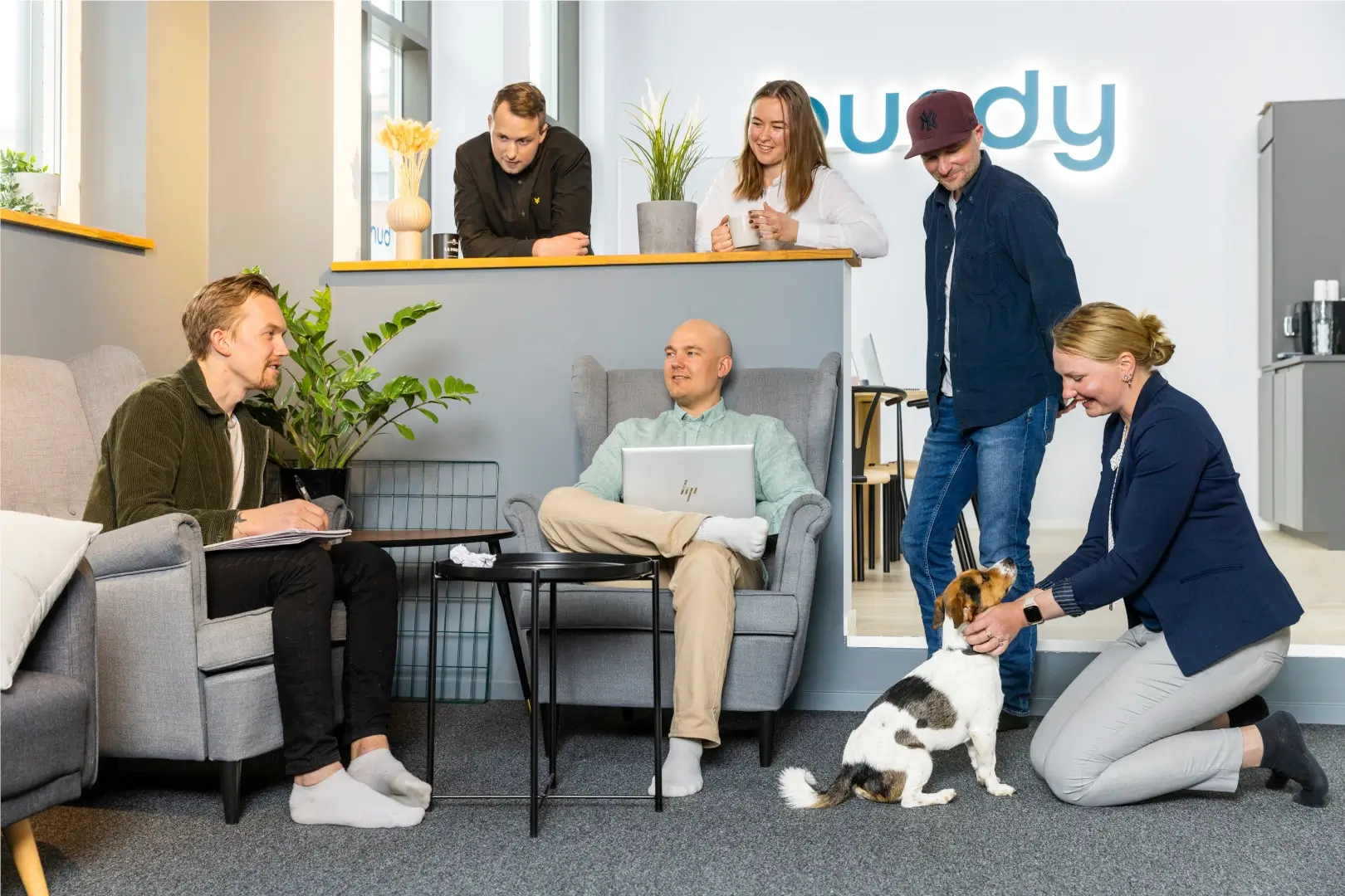The team is sitting and standing together in a nice atmosphere. One of the female colleagues are petting a small dog.